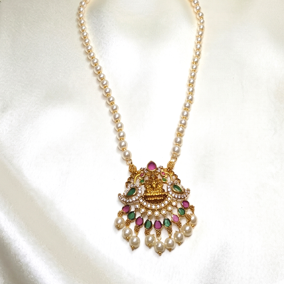 A gold-toned traditional pendant necklace with a pearl chain and a decorative pendant featuring gold metalwork, white pearls, and pink and green gemstone accents, displayed on a white satin fabric background.