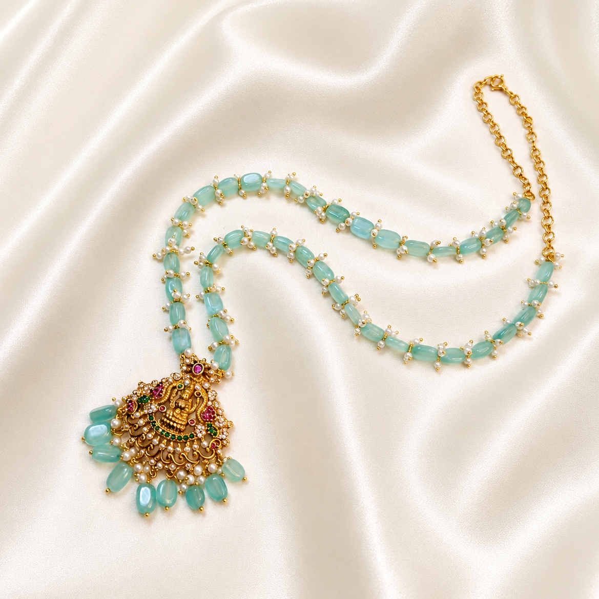 A gold-plated necklace featuring light blue-green teardrop-shaped beads and a detailed pendant with gold, green, and red accents, displayed on a draped cream satin fabric.