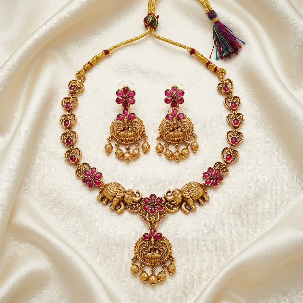 Gold-toned traditional Indian jewelry set featuring a necklace and matching earrings with elephant, floral, and heart motifs, embellished with pink stones, displayed on draped cream satin fabric.