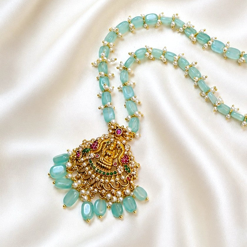 A gold-plated necklace with light green jadeite bead strands and a large ornate gold pendant decorated with faux pearls, green and red stones, and a central red gem, displayed on a white satin background.
