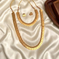 A gold coin necklace set with matching earrings, displayed on draped white satin fabric next to a dark wooden tray. The set includes a long layered coin necklace, a shorter coin necklace, and a pair of small coin earrings.