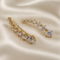 A pair of gold-toned hair clips decorated with a curved row of clear round-cut gemstones, displayed on a draped cream satin fabric background.