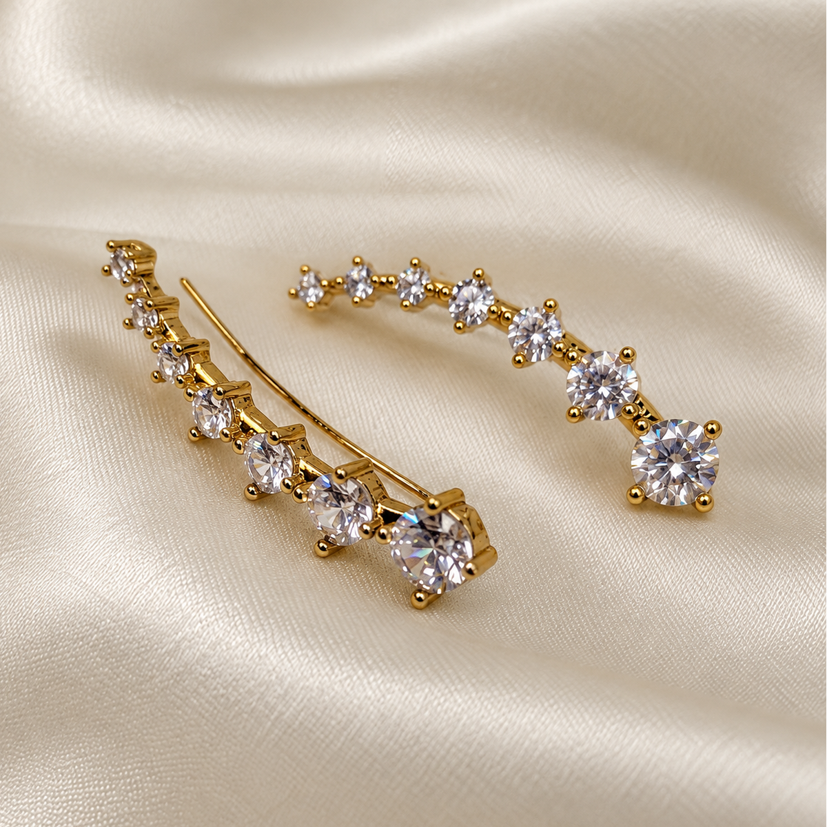 A pair of gold-toned hair clips decorated with a curved row of clear round-cut gemstones, displayed on a draped cream satin fabric background.