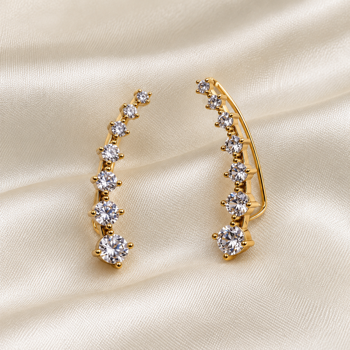 Gold crescent-shaped hoop cuff earrings with a row of clear round-cut cubic zirconia stones, displayed on a draped cream satin fabric.