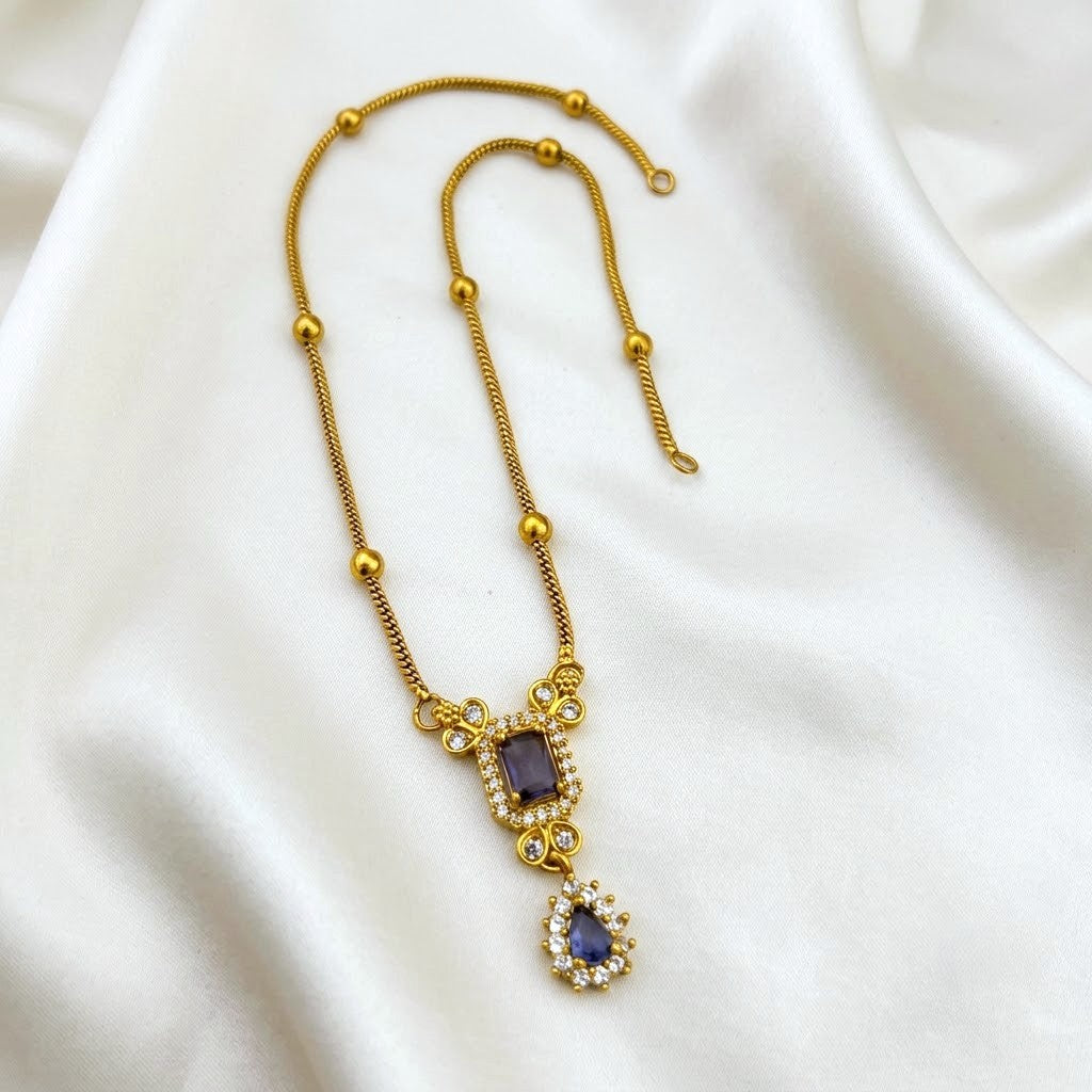 A gold-plated necklace with a textured chain, beaded accents, and a pendant featuring a rectangular purple stone and a teardrop purple stone, accented with small clear crystals, displayed on a white satin fabric background.
