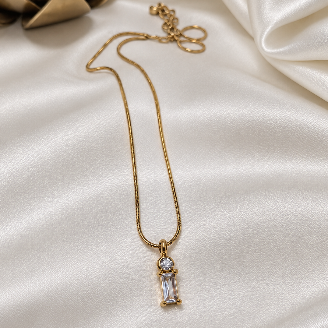 A gold chain necklace with a small rectangular clear gemstone pendant, displayed on draped white satin fabric with a decorative gold object in the corner.