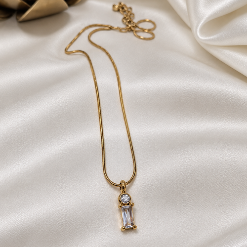 A gold chain necklace with a small rectangular clear gemstone pendant, displayed on draped white satin fabric with a decorative gold object in the corner.