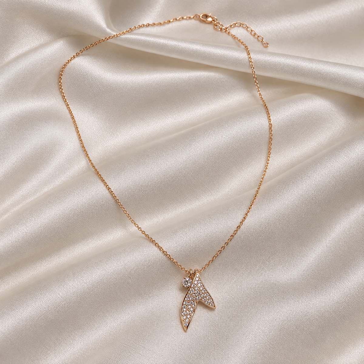 A delicate rose gold chain necklace with a small, leaf-shaped pendant paved with clear crystals, displayed on a draped cream satin fabric background.