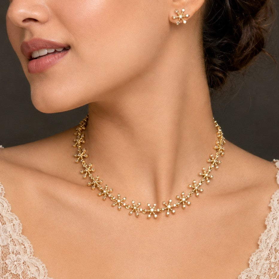 A close-up of a woman wearing a delicate gold necklace and matching stud earring. The jewelry features small, repeating floral or starburst motifs with tiny clear stones set in yellow gold, shown against her neck and upper chest.