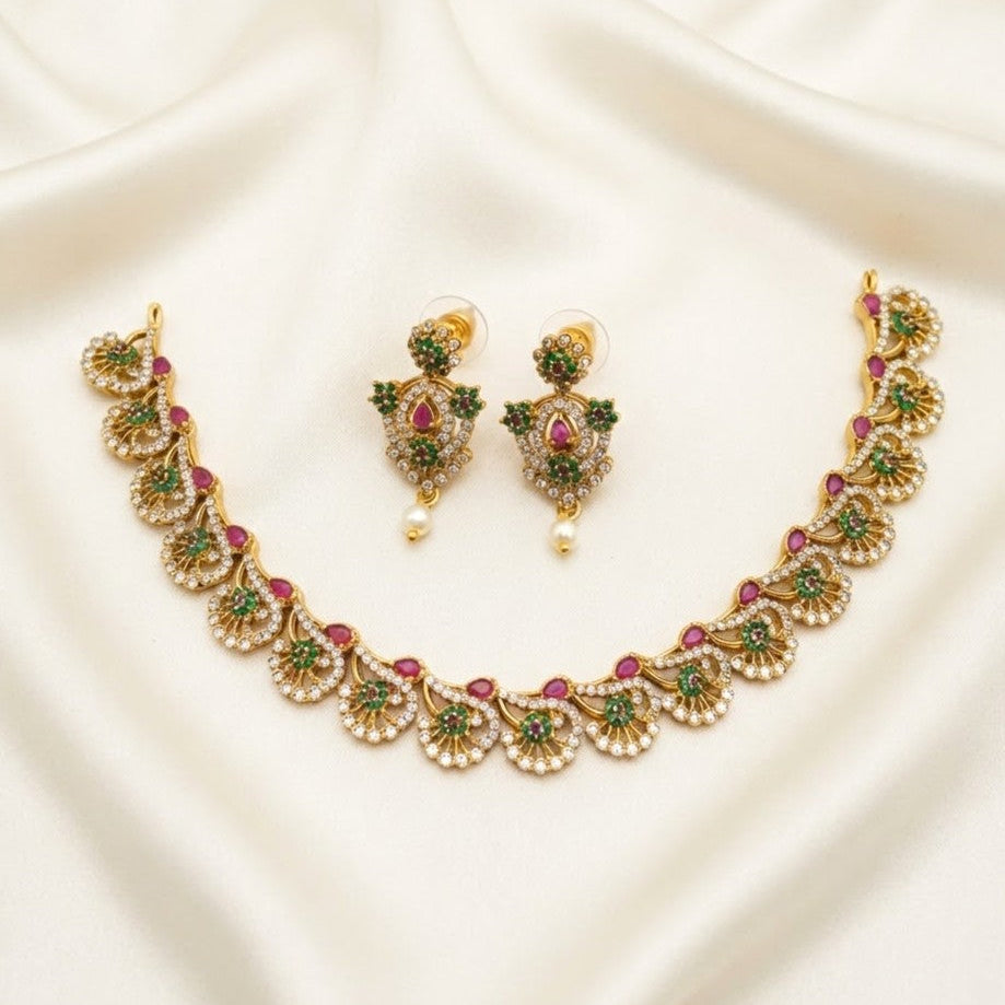Gold-toned traditional Indian jewelry set featuring a necklace and matching earrings, embellished with green stones, pink stones, and white faux pearls, displayed on a draped cream satin fabric.