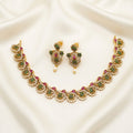 Gold-toned traditional Indian jewelry set featuring a necklace and matching earrings, embellished with green stones, pink stones, and white faux pearls, displayed on a draped cream satin fabric.
