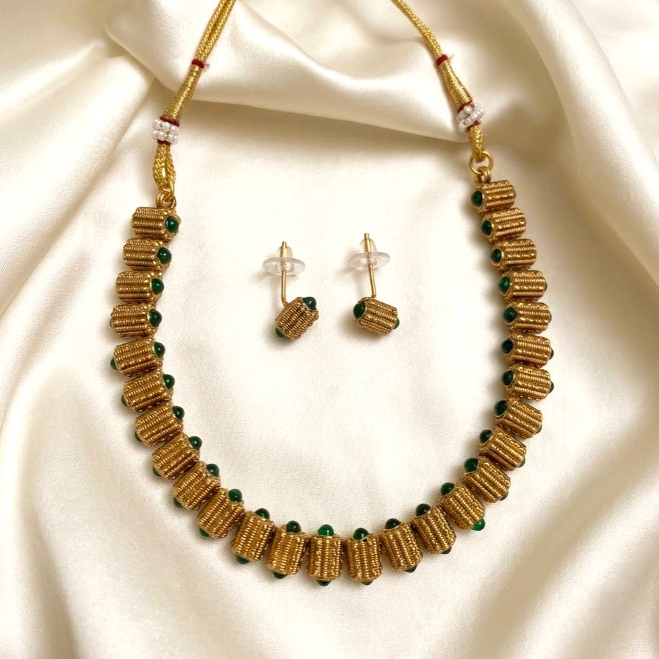 A gold-toned beaded necklace and matching stud earrings laid on draped cream satin fabric. The jewelry features cylindrical textured beads, small dark green stones, and a few pale pink accent beads.