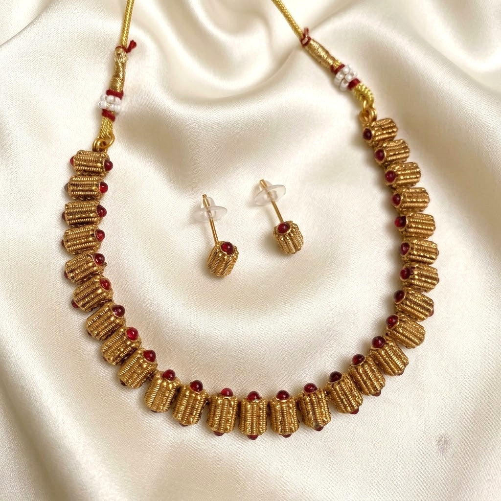 Gold-toned necklace and matching stud earrings with cylindrical textured links and small red bead accents, displayed on a cream satin fabric background.