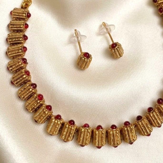 Gold-toned beaded necklace and matching stud earrings with small red gemstone accents, displayed on a cream satin fabric background.