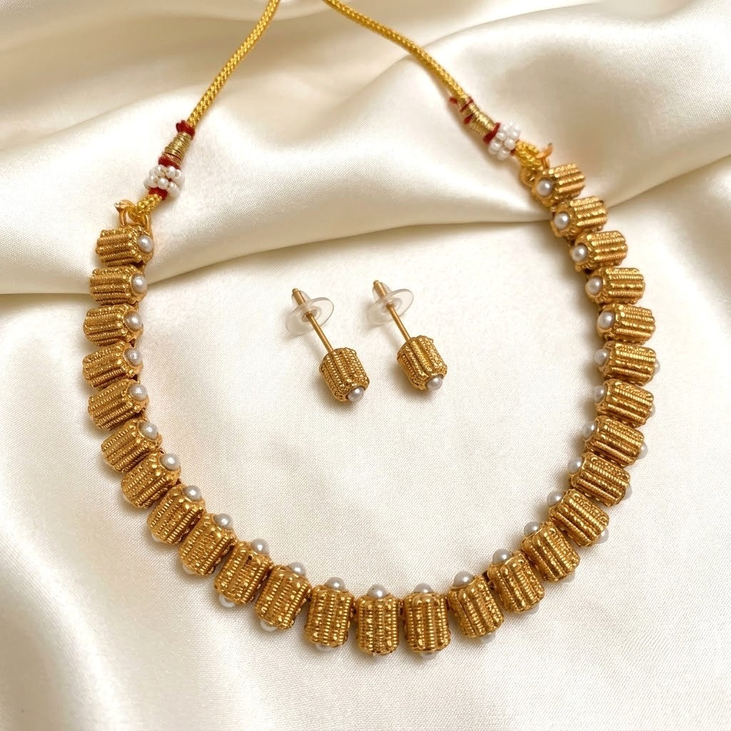 A gold-toned beaded necklace and matching stud earrings displayed on draped cream satin fabric. The necklace features textured cylindrical beads, small white pearl-like accents, and red-and-white spacer details on the cord.