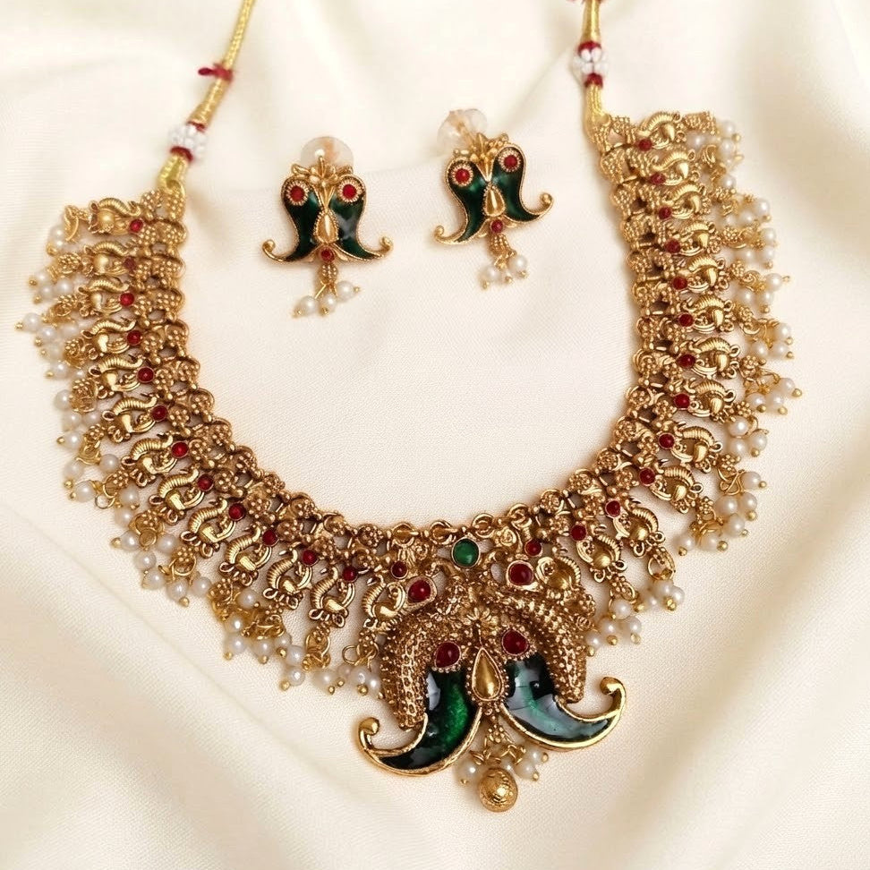 Gold-plated Indian jewelry set featuring a choker necklace and matching earrings with green enamel, red stone accents, and white pearl bead details, displayed on a cream satin fabric background.