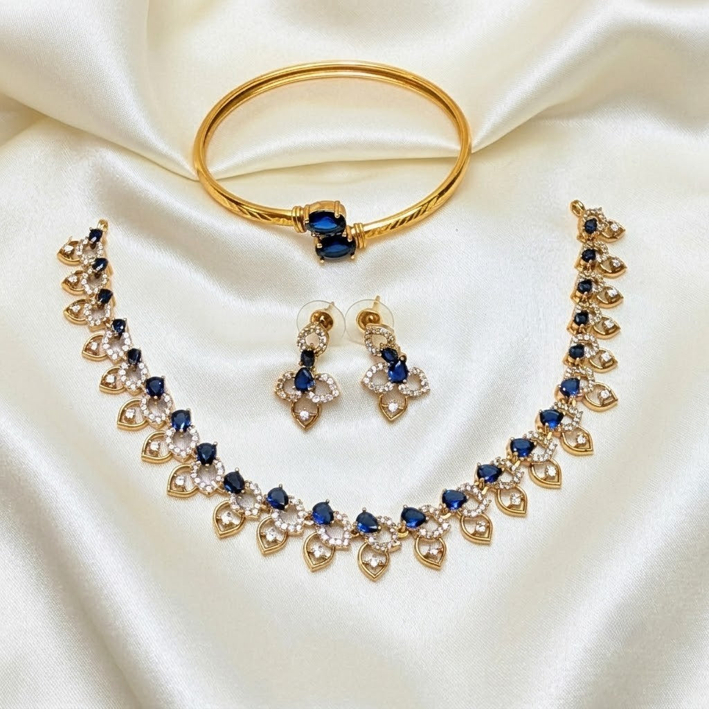 Gold jewelry set with a bangle bracelet, necklace, and matching earrings, featuring deep blue stones and clear crystal accents, displayed on a draped white satin fabric.