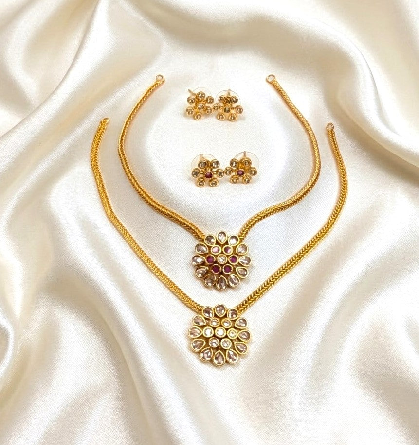 Gold jewelry set featuring a necklace, earrings, and a choker laid out on draped white satin fabric. The floral pendant and matching earrings are decorated with gold and red stone accents.