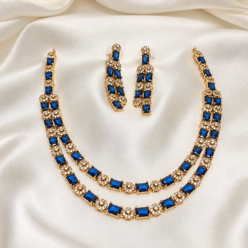 Gold-tone jewelry set with a double-strand necklace, matching drop earrings, and a bracelet, featuring deep blue rectangular stones and clear rhinestone clusters, displayed on a draped white satin fabric.