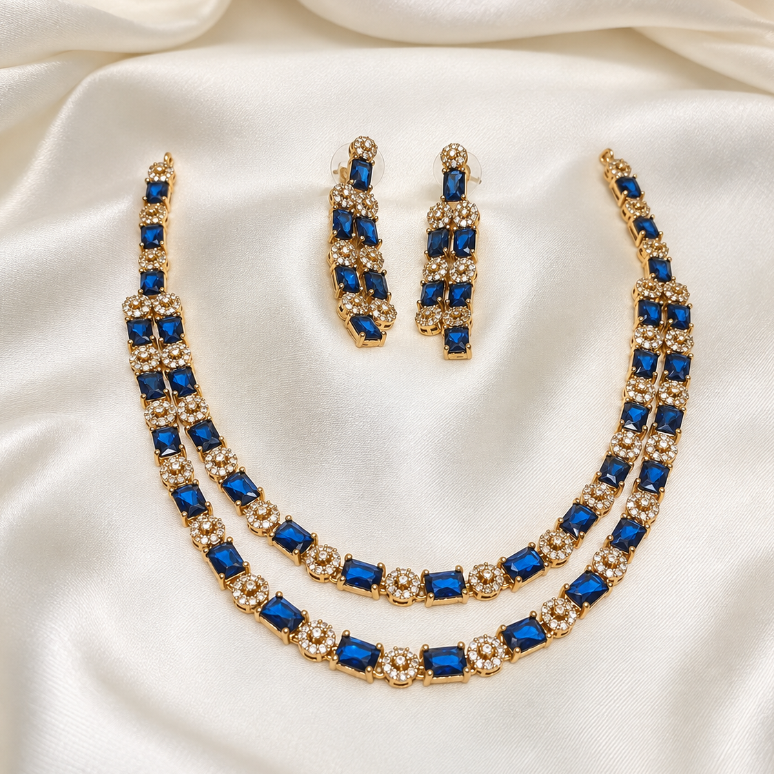 Gold-tone jewelry set with a double-strand necklace, matching drop earrings, and a bracelet, featuring deep blue rectangular stones and clear rhinestone clusters, displayed on a draped white satin fabric.