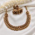 An ornate gold-toned traditional necklace set with matching earrings, arranged on white satin fabric with white flowers at the top. The necklace features multiple rows of engraved round coins and small red and green stone accents.