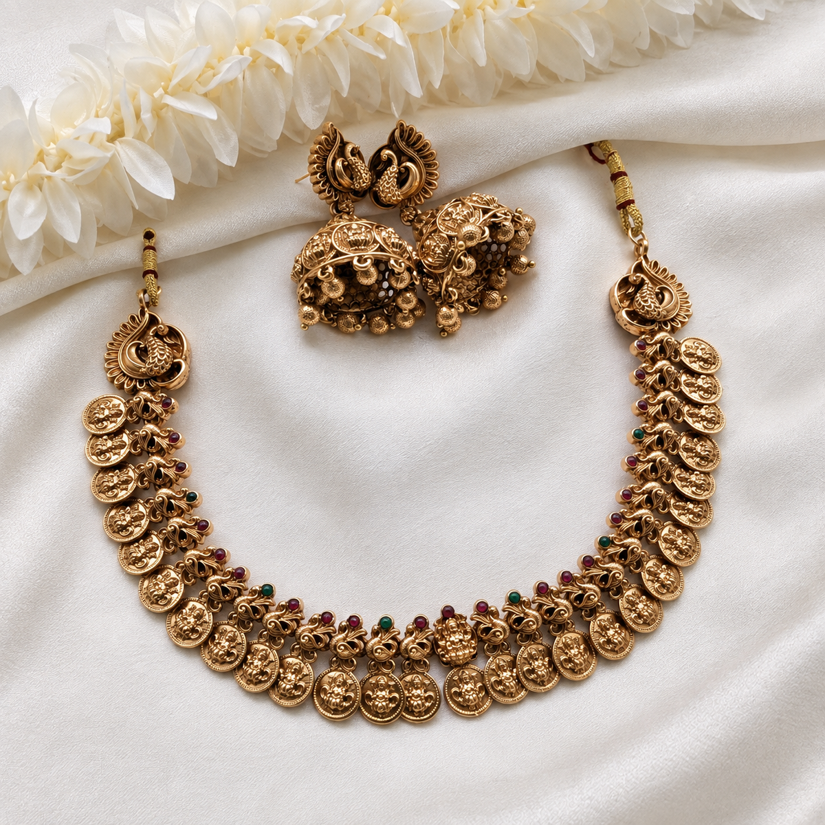An ornate gold-toned traditional necklace set with matching earrings, arranged on white satin fabric with white flowers at the top. The necklace features multiple rows of engraved round coins and small red and green stone accents.