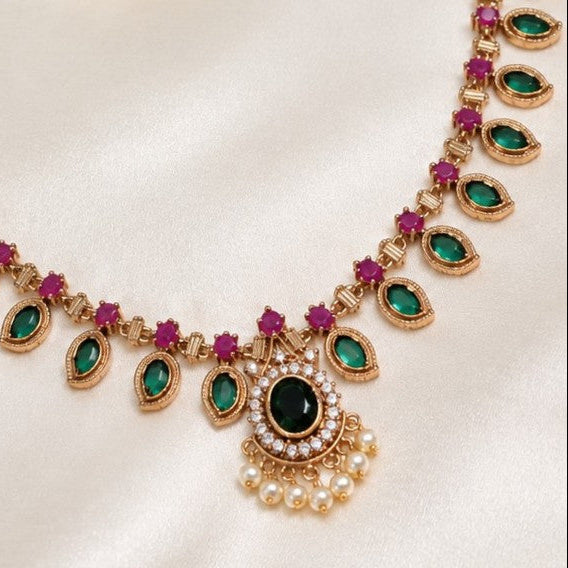 Gold-plated Indian style necklace with green emerald-cut stones, pinkish-red round stones, and a central green oval stone surrounded by white faux pearls, displayed on a cream satin fabric.