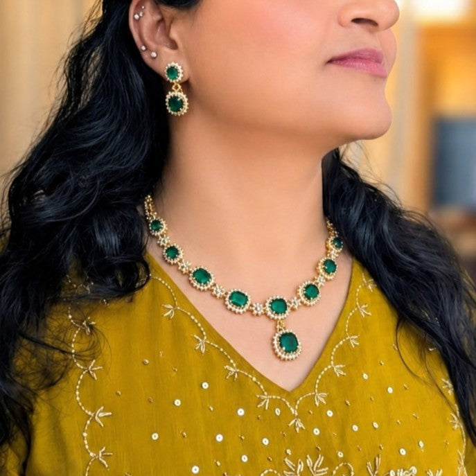 A close-up lifestyle shot of a woman wearing a gold and emerald green jewelry set, featuring a matching necklace and drop earrings, paired with a yellow embroidered traditional top.