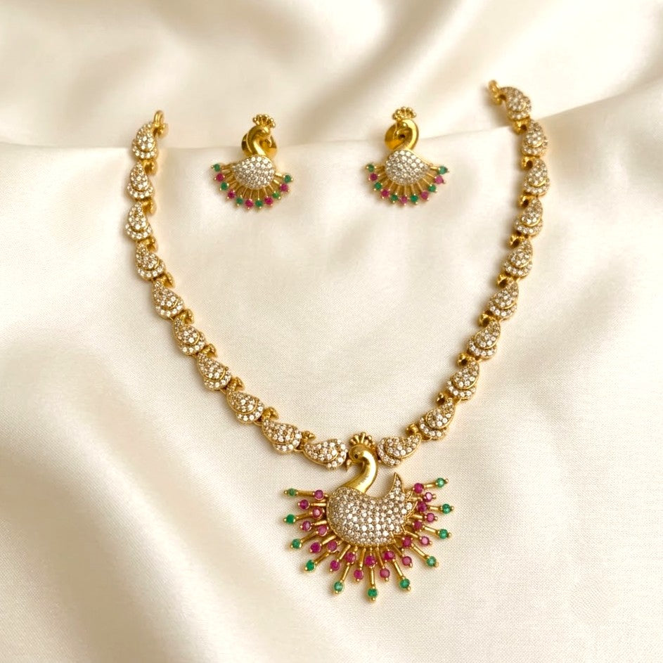Gold-plated Indian jewelry set featuring a peacock-shaped pendant necklace and matching earrings, embellished with clear stones and small green and pink gem accents, displayed on a cream satin fabric background.