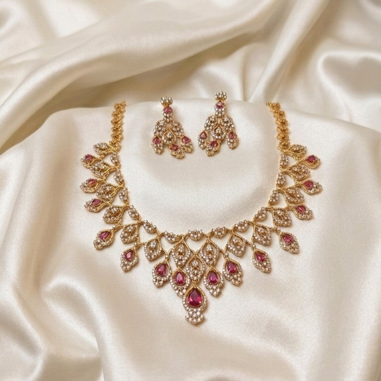 Gold-toned traditional jewelry set featuring a necklace and matching earrings, embellished with small clear stones and pink gemstone accents, displayed on draped cream satin fabric.